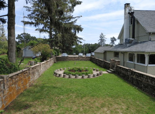 A landscaped yard featuring a circular flower bed surrounded by stone, with a house and trees in the background.