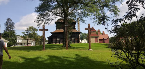 A person walks past three wooden crosses in front of a historic building on a grassy area under a blue sky.