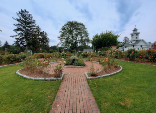A peaceful garden with a circular flower bed, brick path, and trees, set against a cloudy sky and a distant building.