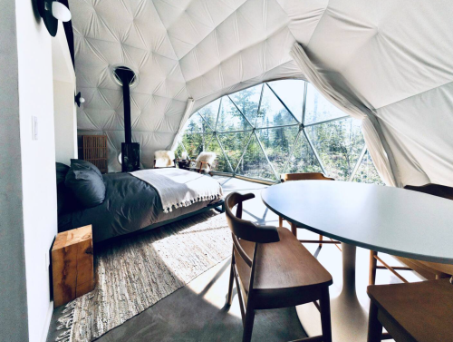 Cozy interior of a geodesic dome with a bed, dining table, and large windows showcasing a natural landscape.