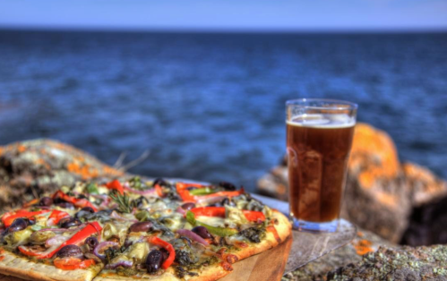 A delicious pizza topped with vegetables sits beside a glass of beer, with a scenic ocean view in the background.