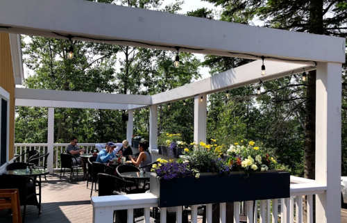 A sunny outdoor patio with people dining and enjoying drinks, surrounded by greenery and flower planters.