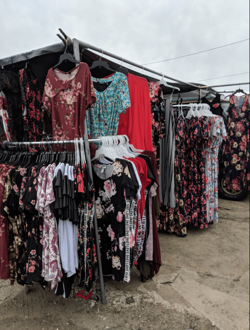 A clothing stall displaying various floral-patterned dresses and tops on hangers, set against a cloudy sky.