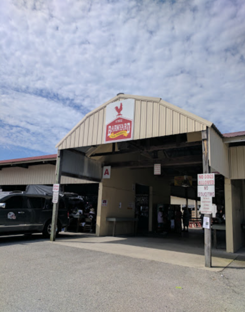 Entrance to a barn-like structure with a sign reading "The Barnyard," under a partly cloudy sky. Vehicles parked nearby.