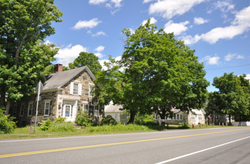 A scenic view of a tree-lined road with stone houses and blue skies dotted with clouds.