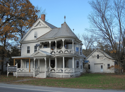 Victorian-style house with a wraparound porch, labeled "Gingerbread Apartments," surrounded by trees and a clear sky.