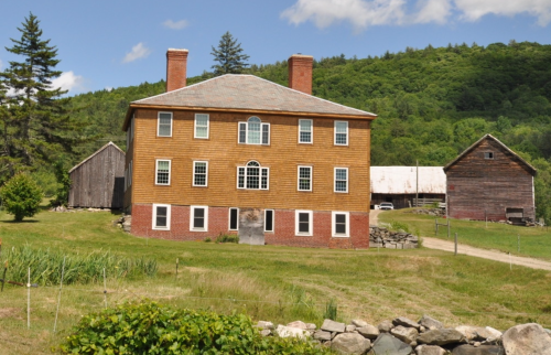 Historic two-story farmhouse with a wooden exterior, surrounded by greenery and a barn in the background.