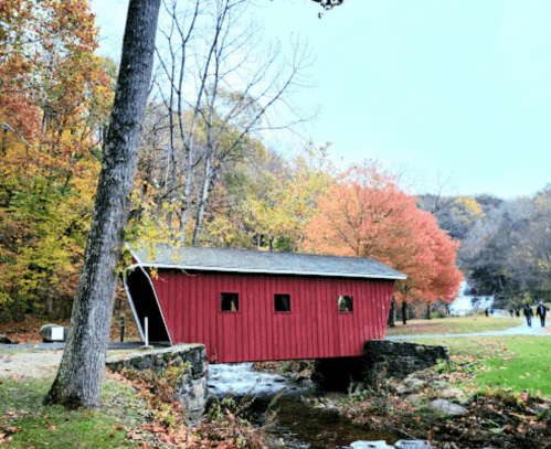 A red covered bridge over a stream, surrounded by autumn trees with vibrant orange and yellow leaves.