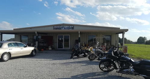 A building with a sign, featuring parked motorcycles and a vintage car in front, set against a grassy field.
