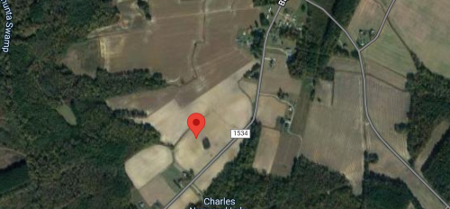 Aerial view of rural land with fields, trees, and a road marked by a red pin indicating a specific location.