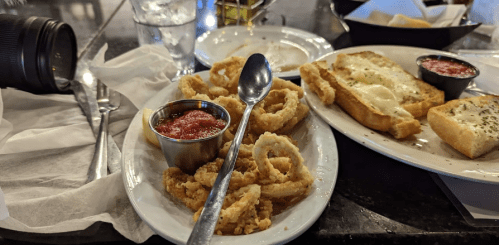A plate of fried calamari with marinara sauce, alongside toasted bread with cheese, on a restaurant table.