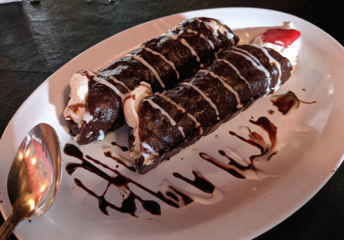 Two chocolate cannoli drizzled with white icing and chocolate sauce, served with whipped cream and a strawberry.