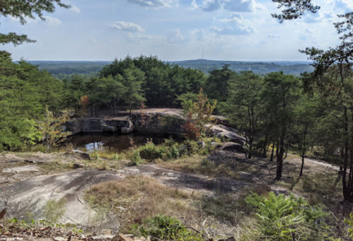 A rocky landscape with trees, a small pond, and distant hills under a partly cloudy sky.