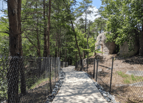 A paved path winds through a wooded area, bordered by a chain-link fence and rocky outcrops under a clear blue sky.