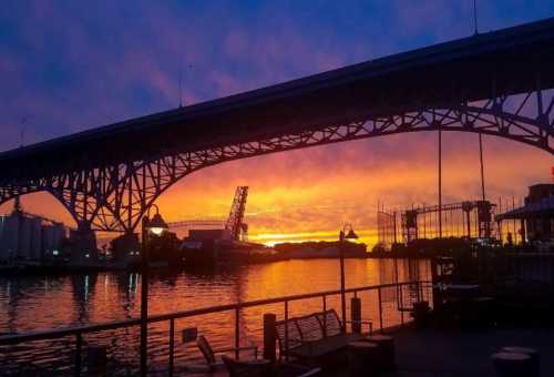 A vibrant sunset casts colorful hues over a river, framed by a large bridge and silhouettes of nearby structures.