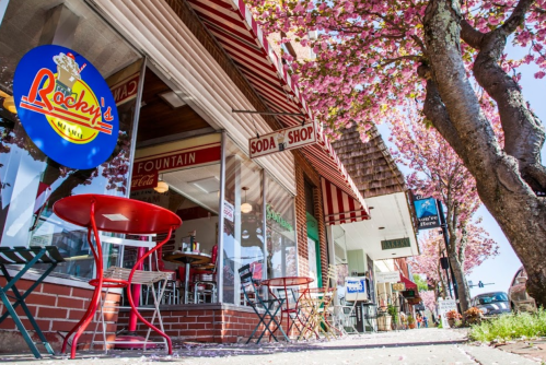 A charming soda shop with outdoor seating, surrounded by blooming cherry trees and colorful storefronts.