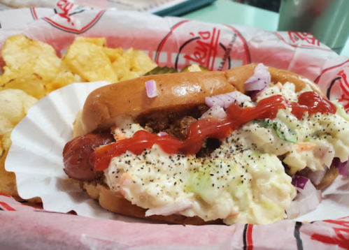 A hot dog topped with coleslaw, onions, and ketchup, served with a side of potato chips on a paper tray.