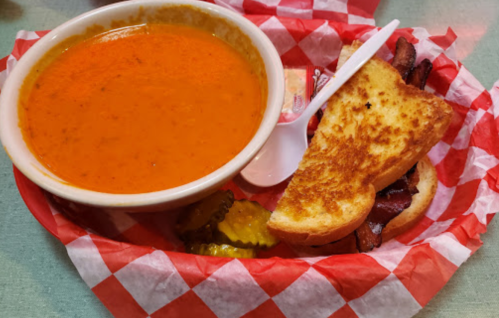 A bowl of tomato soup with a grilled cheese sandwich, pickles, and a butter packet on a red-checkered tray.