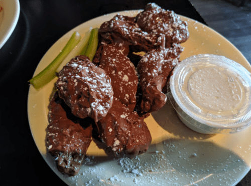 A plate of chocolate-covered chicken wings dusted with powdered sugar, served with a side of dipping sauce and celery sticks.