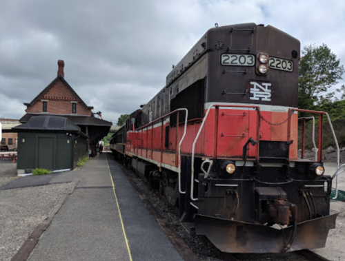 A vintage train engine parked at a station, with a historic building in the background under a cloudy sky.