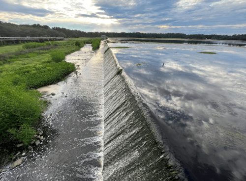 A serene landscape featuring a dam with water cascading over, surrounded by lush greenery and a cloudy sky.