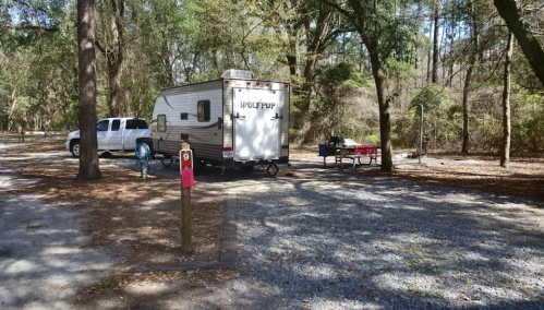 A camper parked at a campsite with a truck nearby, surrounded by trees and gravel. A picnic table is visible.