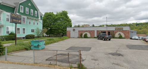 A parking lot with a gray building labeled "Grotto" and a green dumpster, next to a large green house.