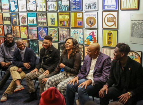 A group of six people sitting together, smiling and laughing, in front of a wall of colorful music album covers.