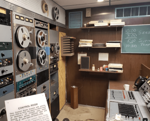 Vintage control room with reel-to-reel tape machines, a chalkboard, and various audio equipment on wooden walls.