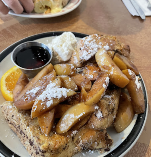 A plate of French toast topped with sautéed apples, powdered sugar, a dollop of whipped cream, and a side of syrup.