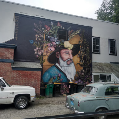 A colorful mural of a bearded man in a hat surrounded by flowers, on a building next to vintage cars and trash bins.