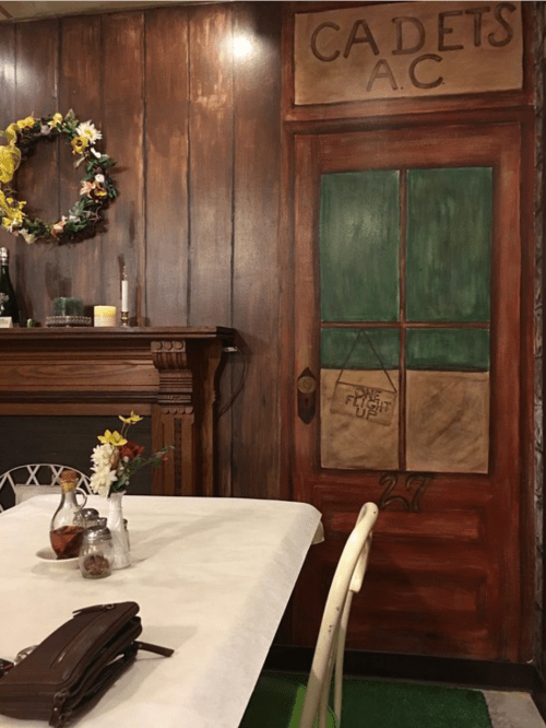 A cozy restaurant interior featuring a wooden wall, a painted door labeled "CADETS A.C.," and a table with flowers.