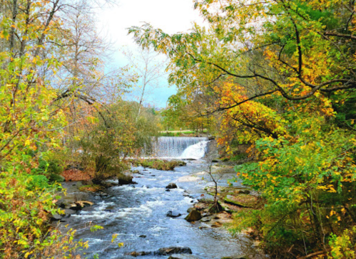 A serene river flows past colorful autumn trees, with a small waterfall in the background under a cloudy sky.