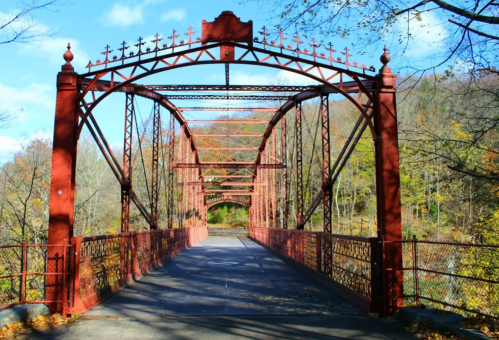 A red, ornate metal bridge spans a river, surrounded by colorful autumn foliage and a clear blue sky.