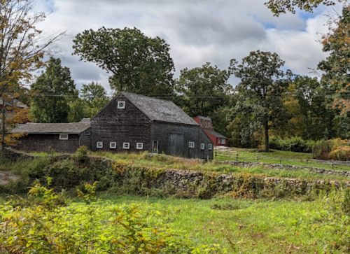 A rustic barn surrounded by greenery and trees under a cloudy sky, with a stone wall in the foreground.