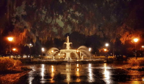 A illuminated fountain in a park at night, surrounded by trees and wet pavement, with water gently flowing.