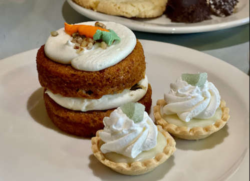 A layered carrot cake with cream cheese frosting, topped with nuts, alongside two mini lemon tarts with whipped cream.