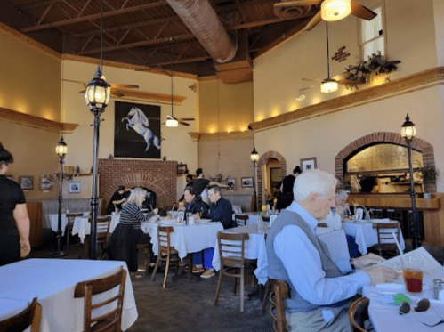 A cozy restaurant interior with diners at tables, warm lighting, and a large painting of a horse on the wall.