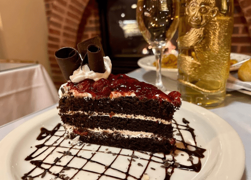 A slice of chocolate cake topped with cherries, whipped cream, and chocolate curls, served on a white plate.