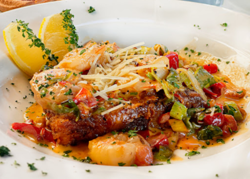 A plate of seafood topped with vegetables, cheese, and garnished with lemon wedges and herbs.