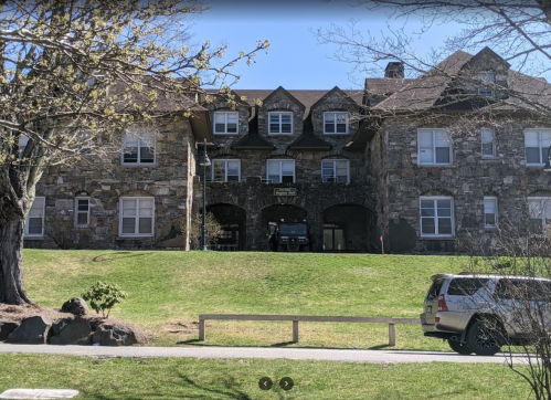 Stone building with multiple windows and arches, set on a grassy area with a tree and a parked vehicle in front.
