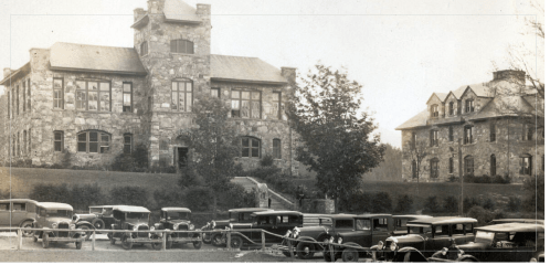Historic stone buildings with large windows, surrounded by vintage cars parked in front.