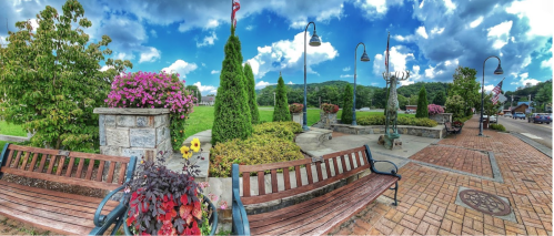 A scenic park with benches, colorful flowers, trees, and a statue, under a bright blue sky with fluffy clouds.