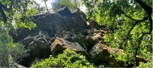A rocky cliff surrounded by lush green trees and foliage under a bright blue sky.