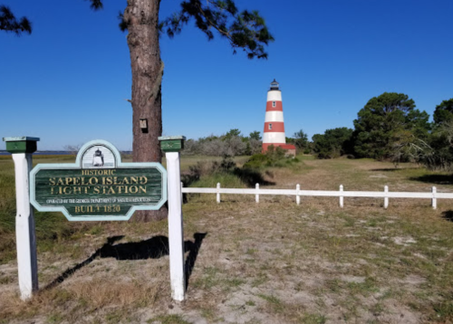 A sign for Sapelo Island Light Station with a red and white lighthouse in the background against a clear blue sky.