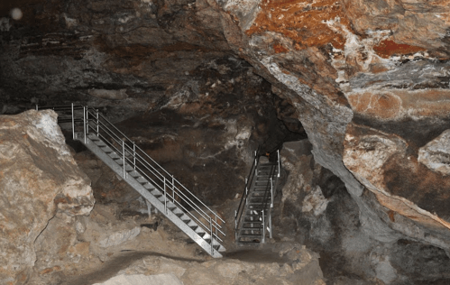 Metal stairs lead up through a rocky cave, surrounded by textured stone walls and earthy ground.