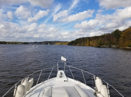 View from the bow of a boat on a river, surrounded by trees with autumn foliage under a cloudy sky.