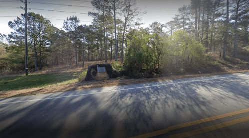 A roadside view featuring trees and a stone monument partially obscured by bushes.
