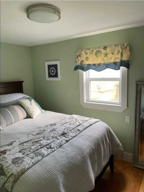 Cozy bedroom with a green wall, a bed with light bedding, a window with a patterned valance, and a mirror.