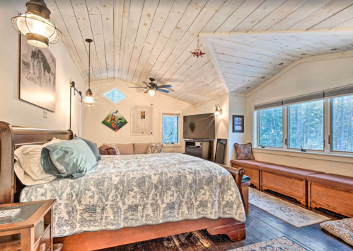 Cozy bedroom with a wooden ceiling, large bed, and windows overlooking a forested area. Warm, inviting decor.